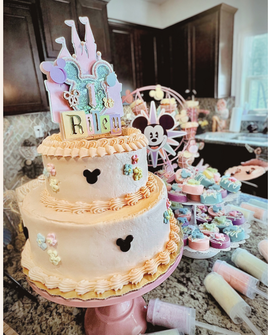 A pastel Disney-themed birthday cake with “1 Riley” topper sits on a pink stand, surrounded by Mickey-shaped cookies, cupcakes, and cotton candy push pops. The setup features soft pastel colors, a Mickey Ferris wheel, and kitchen counter decor.