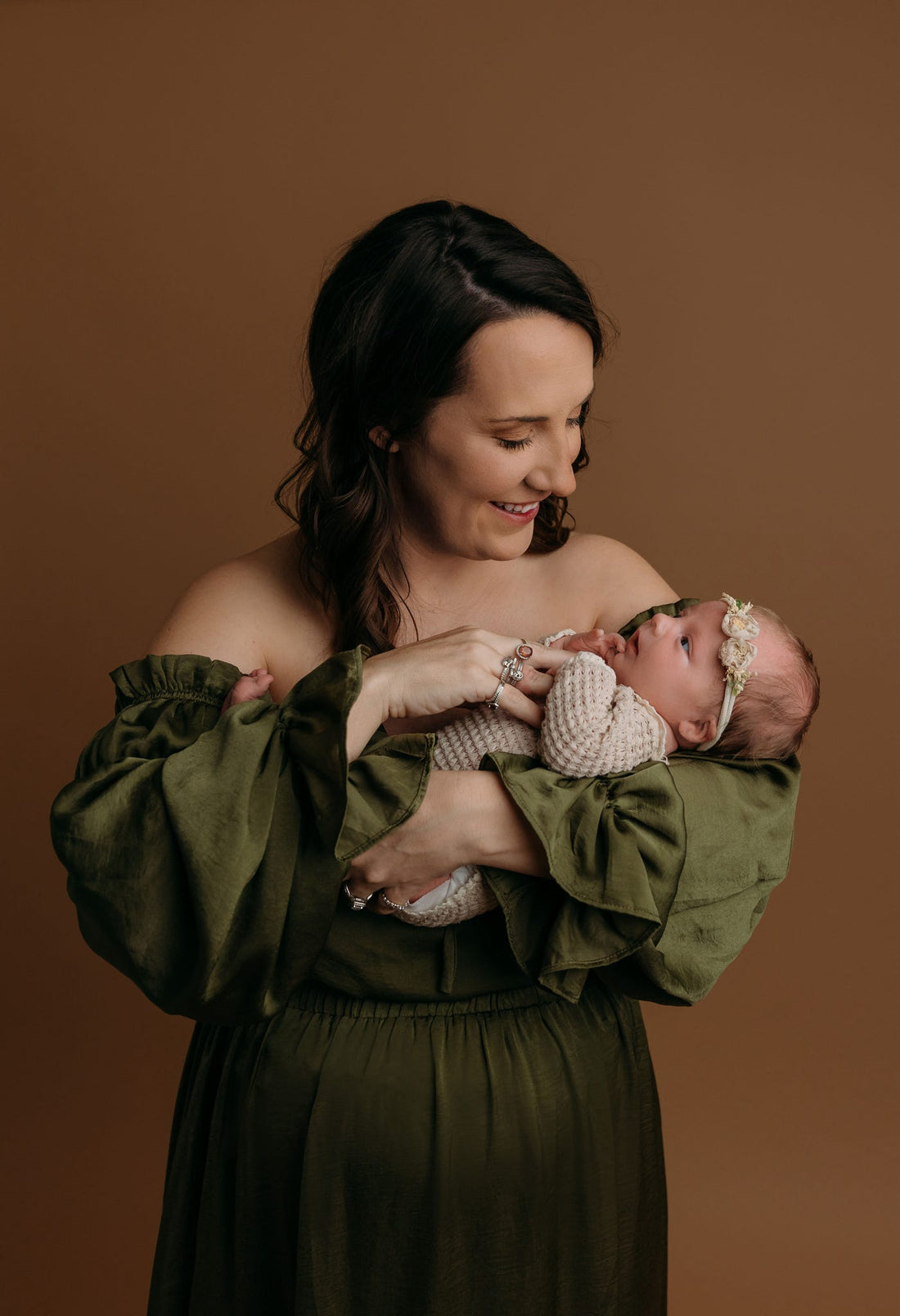 A mother in an off-the-shoulder olive green dress smiles while holding her newborn baby against a warm brown backdrop. The baby wears a soft knit outfit and a floral headband. The moment captures tender connection and maternal love in a studio setting.