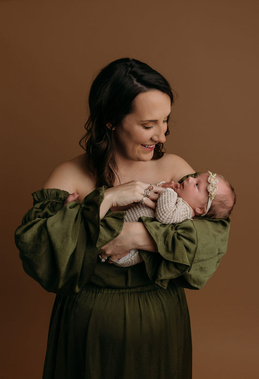A mother in an off-the-shoulder olive green dress smiles while holding her newborn baby against a warm brown backdrop. The baby wears a soft knit outfit and a floral headband. The moment captures tender connection and maternal love in a studio setting.