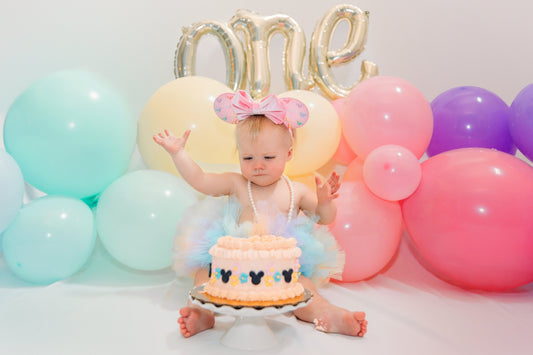 A baby in a pastel tutu and pink Minnie ears sits barefoot in front of a peach-colored smash cake with Mickey silhouettes. Surrounded by pastel balloons and a light gold “one” balloon, she raises one hand mid-smash, celebrating her first birthday.