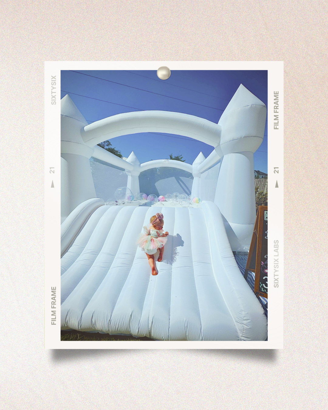 Toddler in a pastel tutu climbs a large white inflatable bounce house under a clear blue sky, with colorful balloons at the top.