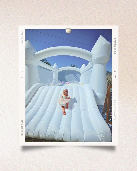 Toddler in a pastel tutu climbs a large white inflatable bounce house under a clear blue sky, with colorful balloons at the top.