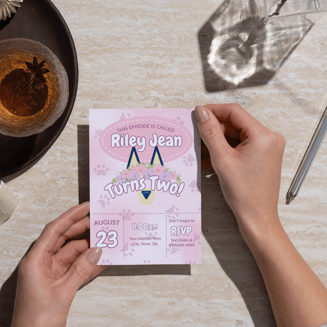 A person holds a pastel pink Bluey-inspired birthday invitation for a little girl named Riley Jean. The design features dog ears with a floral crown and paw prints. Text reads “Riley Jean Turns Two!” with party details and RSVP info below.