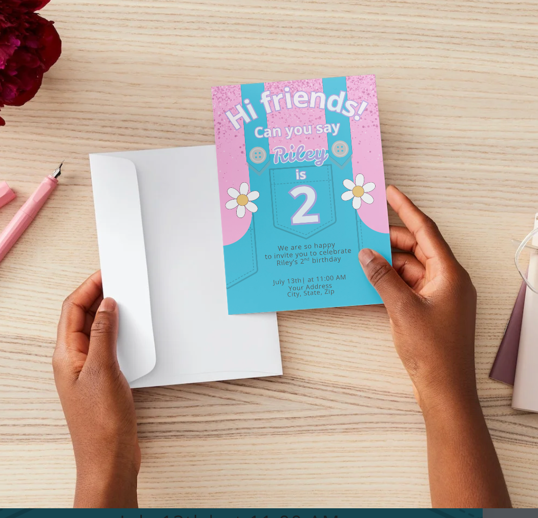Hands holding a Mrs. Rachel-inspired second birthday invitation featuring pink glitter, blue overalls, daisies, and playful fonts. Text reads “Hi friends! Can you say Riley is 2.” Shown above a white envelope on a light wood desk with pink pens.