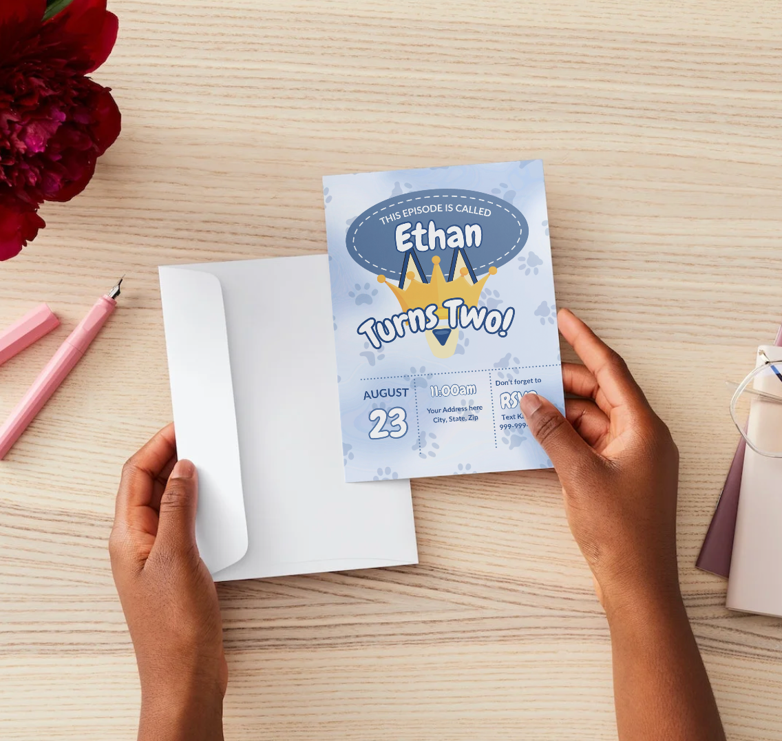 A person holds a Bluey-inspired second birthday invitation that reads “This episode is called Ethan Turns Two!” above a paw print background with a gold crown illustration. The invite is shown over an open white envelope on a light wood surface.