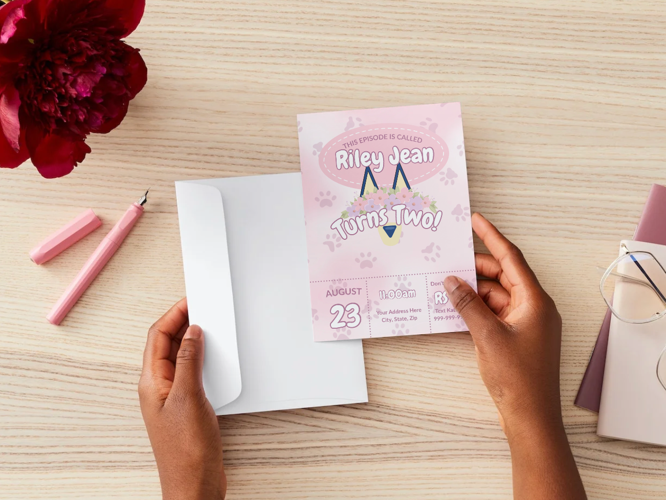 A person holds a pastel pink Bluey-inspired birthday invitation over a wooden table. The invite reads “This Episode Is Called Riley Jean Turns Two!” with floral accents, paw prints, and event details for August 23 at 11:00 AM.